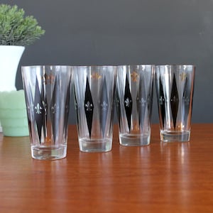 May include: Four vintage clear drinking glasses with black diamond and fleur-de-lis designs. The glasses are arranged on a wooden surface, with decorative items in the background.