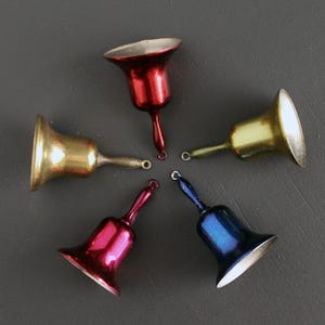 Peut inclure: Cinq petites cloches décoratives de couleurs variées, disposées en cercle. Les cloches sont dorées, rouges, roses, bleues et jaunes. Chaque cloche a une petite boucle et un intérieur argenté.