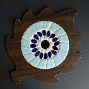 Puede incluir: Una base de madera marrón oscuro con forma de hoja, que presenta una incrustación de mosaico circular. El mosaico consta de un orificio central negro rodeado de azulejos azul oscuro en forma de pétalo, un anillo blanco y un borde azul claro.