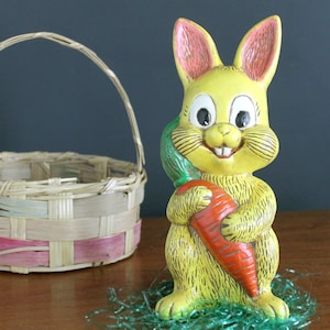 May include: A yellow ceramic Easter bunny figurine holding a large orange carrot, with a green leafy base. A woven basket with pink and light blue accents sits to the left. The bunny has pink inner ears and a smiling face.
