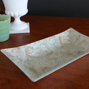 May include: Rectangular, light green glass tray with gold floral detailing. The tray has a slightly raised edge and is set on a wooden surface. A jadeite green glass cup and a white vase are also in the image.