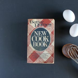 May include: A vintage cookbook titled "New Cook Book" with a red and white checkered pattern cover. Two white eggs and a wooden whisk are next to the book on a dark gray surface. The cookbook has the words "Better Homes" on the top.