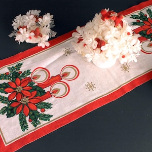 May include: A white table runner with a red border, featuring a Christmas poinsettia and candle design. The runner is decorated with gold snowflakes and is paired with white floral arrangements.