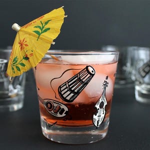 May include: A cocktail glass filled with a pink drink and ice cubes, garnished with a yellow paper umbrella. The glass is decorated with black and white illustrations of musical instruments. Several similar glasses are in the background.