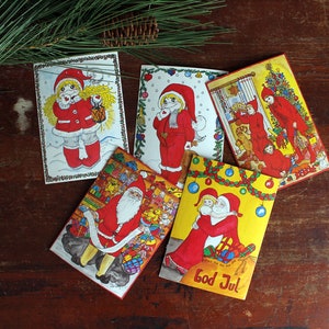 May include: A collection of vintage Christmas cards featuring illustrations of Santa Claus and festive characters in red and white attire. The cards are arranged on a dark wooden surface, with pine needles and a pine cone in the background.