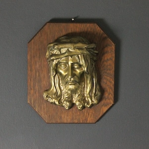 May include: A brass relief of a man's face, with long hair and a beard, wearing a crown of thorns. The relief is mounted on a dark brown, octagonal wooden plaque. Religious art.