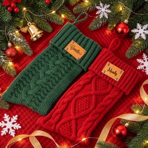 May include: Two knitted Christmas stockings, one dark green and one red, featuring a cable knit pattern. Each stocking has a rectangular tan leather tag attached to the cuff, with the name Camille on the green stocking and Mandy on the red one.