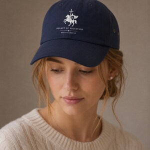 May include: A navy blue baseball cap featuring a white embroidered graphic of a knight on horseback holding a cross-topped staff. Below the graphic, the text reads "HELMET OF SALVATION" followed by a small cross and "REVIVAL HOUSE" in white.