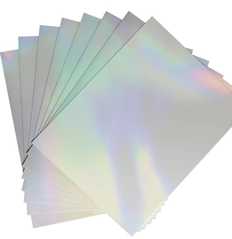 May include: A stack of rectangular sheets of paper with a smooth, reflective surface that displays a holographic, iridescent effect. The sheets are layered in a fan-like arrangement, showing a spectrum of light colors against a silver base.