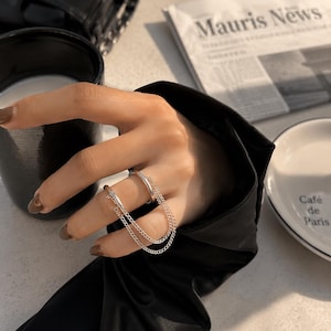 Peut inclure: Bague double doigt argentée avec deux anneaux reliés et de fines chaînes. La bague est portée sur une main aux ongles manucurés. Une tasse noire et un journal avec le texte "Mauris News" sont en arrière-plan.