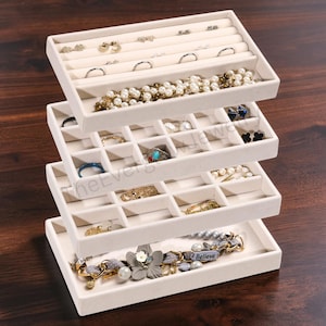 May include: A four-tiered stackable jewelry organizer tray set in a light beige velvet finish. The top tray features ring slots, while the lower three trays are divided into square compartments containing various bracelets, rings, and earrings.