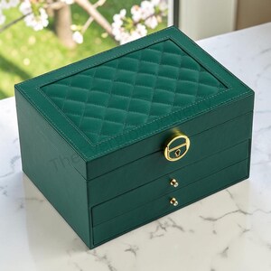 May include: A rectangular dark green jewelry box with a quilted top panel. The box features a gold-toned circular latch on the lid and two lower drawers, each equipped with a small, clear, crystal-like knob for opening the storage compartments.