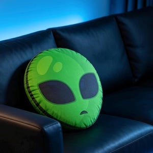 Peut inclure: Un coussin rond vert représentant un visage d'extraterrestre avec de grands yeux noirs et une petite bouche. Le coussin est posé sur un canapé en cuir noir. Le visage extraterrestre est vert clair avec un contour vert plus foncé.