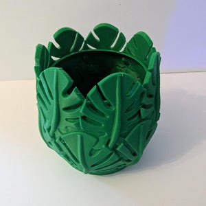 May include: A vibrant green cylindrical planter featuring a repeating relief pattern of monstera leaves covering the entire exterior surface. The top edge is scalloped, following the individual shapes of the leaves, creating a textured design.
