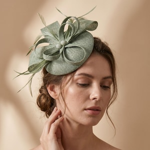 Peut inclure: Un fascinateur vert sauge, en tissu texturé, orné d'un grand nœud et de plumes délicates. Le couvre-chef présente un ornement central et est conçu pour les occasions spéciales.
