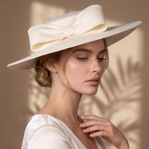 Può includere: Un cappello a tesa larga color crema caratterizzato da una corona strutturata e un grande fiocco prominente avvolto attorno alla base. Il cappello è realizzato in un materiale liscio e solido con una finitura pulita e una tesa larga.