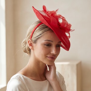 Könnte beinhalten: Ein leuchtend rotes Kopfstück mit einem Stirnband und einem geschichteten Design. Das Kopfstück hat eine breite Krempe und ist mit Federn verziert. Dieses Accessoire eignet sich für besondere Anlässe wie Hochzeiten oder formelle Veranstaltungen.