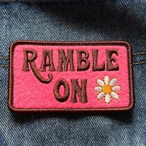 Könnte beinhalten: Ein pinkfarbener rechteckiger Aufnäher mit dem Schriftzug "RAMBLE ON" in brauner Schrift und einer kleinen gestickten Gänseblümchen. Der Aufnäher ist auf der Gesäßtasche einer blauen Jeans aufgenäht.