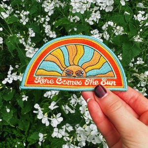 May include: Embroidered patch with a sun design in a half-circle shape. The patch features blue, orange, and yellow colours, with the text "Here Comes The Sun" in white. The patch is held against a backdrop of white flowers and green foliage.