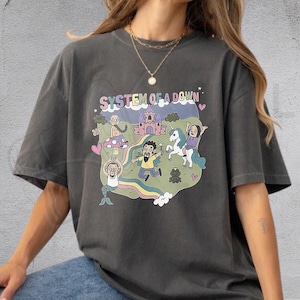Op de afbeelding: Een antracietgrijs T-shirt met korte mouwen en een kleurrijke grafische print met de tekst "SYSTEM OF A DOWN". De illustratie toont een kasteel, een eenhoorn, een zeemeermin en diverse figuren in een groen landschap met een regenboog.