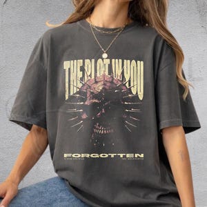 以下が含まれることがあります： ダークグレーの半袖Tシャツで、暗いスパイク付きの頭蓋骨のグラフィックプリントが施されています。頭蓋骨の上にはかすれたベージュのフォントで「THE PLOT IN YOU」という文字があり、グラフィックの下にはベージュで「FORGOTTEN」とプリントされています。