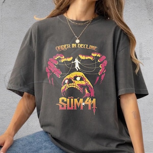 May include: A charcoal gray short-sleeve t-shirt featuring a graphic print of a yellow skull with pink skeletal hands manipulating strings above it. The text reads ORDER IN DECLINE at the top and SUM 41 at the bottom in a stylized font.
