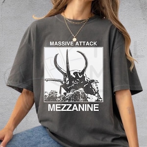 May include: A charcoal gray short-sleeve t-shirt featuring a white square graphic print. The graphic displays a black silhouette of a beetle with large horns, with the text MASSIVE ATTACK above and MEZZANINE below the square in white lettering.