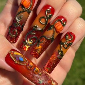 May include: Long square-shaped artificial nails featuring a red and orange gradient base with gold glitter. Three nails are decorated with raised 3D orange pumpkins and metallic gold vines. One nail features faceted iridescent rhinestones and leaf accents.