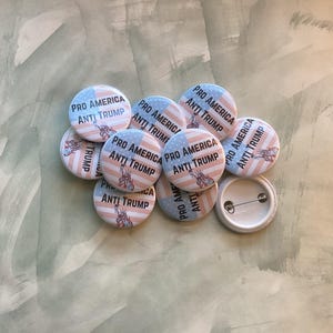 Puede incluir: Un conjunto de botones redondos con un diseño patriótico. Cada botón presenta un fondo de bandera estadounidense estilizado con el texto "PRO AMERICA ANTI TRUMP". Los botones son blancos con un respaldo de alfiler.