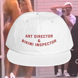 May include: A white baseball cap featuring the red text ART DIRECTOR & BIKINI INSPECTOR printed on the front panel. The cap has a structured crown, a flat brim, and a button on the top center. The design is centered on the front of the hat.
