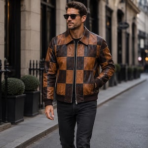 May include: A leather jacket featuring a brown and dark brown checkered pattern, a classic collar, a full-length front zipper, and ribbed cuffs. The jacket is worn over a black shirt and paired with dark gray jeans, complemented by black sunglasses.
