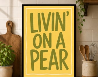 Könnte beinhalten: Ein rechteckiger Wandkunstdruck mit einem hellgelben Hintergrund, einem dünnen weißen Rand und dem dunkel olivgrünen Text "LIVIN' ON A PEAR" in einem kräftigen, handgeschriebenen Stil. Der Druck ist in einem einfachen schwarzen Rahmen gefasst.