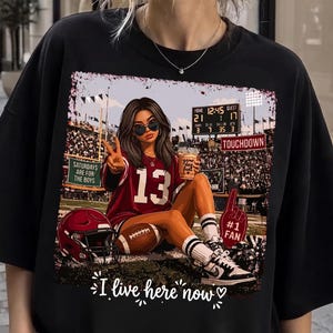 Puede incluir: Una camiseta negra con el gráfico de una persona con una camiseta color granate con el número 13, sentada en un campo de fútbol americano. El gráfico incluye un marcador, un casco, un dedo de espuma y el texto: SATURDAYS ARE FOR THE BOYS, TOUCHDOWN e I live here now.