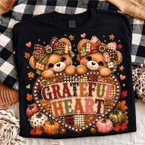 May include: A black t-shirt featuring two brown teddy bears with patterned bows on their heads, positioned above a large heart-shaped graphic containing the text "GRATEFUL HEART" surrounded by various pumpkins and autumn leaves in warm colors.