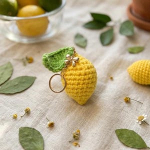 Pode incluir: Um porta-chaves de limão em crochê amarelo com uma superfície texturizada, uma folha de crochê verde presa no topo e um anel de metal dourado com um pequeno pendente decorativo em forma de laço. O limão é feito com pontos densos.