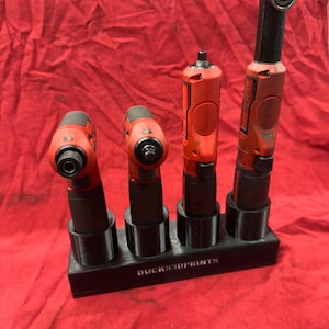 May include: A black plastic tool organizer stand with four circular slots holding four red and black power tools. The base of the stand features the white text DUCKS3DPRINTS. The tools are arranged in a row, with the tallest tool on the far right.