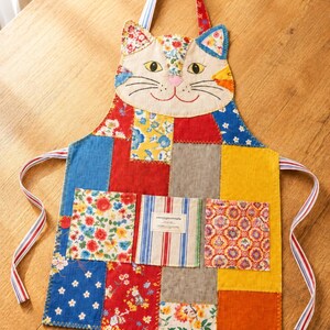 May include: A patchwork apron featuring a cat-shaped top with embroidered facial features. The body consists of various colorful fabric squares in red, blue, yellow, and grey, including floral and striped patterns. It has three front pockets and striped ties.