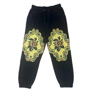 May include: Black sweatpants with a drawstring waist and elastic cuffs. The sweatpants feature a yellow floral design on each leg. The floral design includes flowers in shades of brown, green, and yellow.