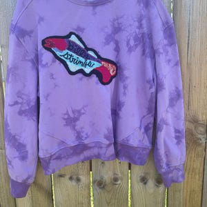 Peut inclure: Un sweat-shirt à col rond et manches longues avec un motif tie-dye violet. L'avant présente un grand écusson brodé représentant un poisson avec le texte "Still Strings" cousu en fil bleu sur le corps du poisson, qui comporte des accents rouges et blancs.
