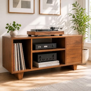Peut inclure: Un meuble multimédia en bois de style moderne du milieu du siècle avec un tourne-disque sur le dessus, un compartiment de rangement vertical pour vinyles, deux étagères ouvertes avec équipement audio et livres, et une porte fermée sur quatre pieds.