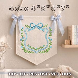 May include: A cream-colored fabric wall banner featuring an embroidered blue shield frame, green vines, and light blue flowers. The banner is suspended by a rope and adorned with two light blue satin bows. Text reads: 4 size 4" 5" 6" 7" EXP - JEF - PES - DST - VP3 - HUS.