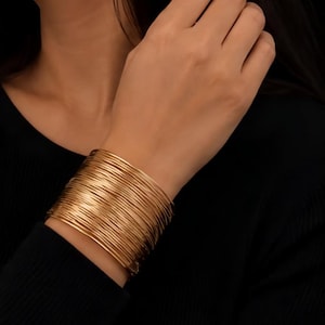 Puede incluir: Una pulsera de brazalete ancho compuesta por numerosos anillos de alambre fino de tono dorado apilados. El accesorio presenta un diseño continuo y superpuesto que crea una apariencia metálica texturizada en la muñeca con acabado pulido.