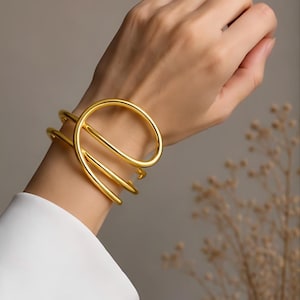May include: A gold-toned cuff bracelet featuring a continuous, multi-layered wire design that wraps around the wrist. The metal has a smooth, polished finish with a rounded profile, creating a series of overlapping circular loops.