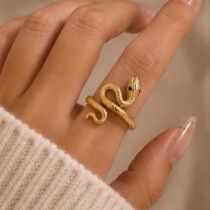 Op de afbeelding: Een goudkleurige metalen ring in de vorm van een kronkelende slang. De ring heeft een textuur die op schubben lijkt over het hele lichaam van de slang en twee kleine, donkere, ronde accenten die de ogen op de kop van de slang voorstellen.