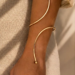 Op de afbeelding: Een goudkleurige armmanchet met een slank, open spiraalontwerp dat om de onderarm wikkelt. Het sieraad is vervaardigd uit een dunne, gepolijste metaaldraad en eindigt aan beide uiteinden in kleine, gladde, bolvormige kralen voor een strakke afwerking.