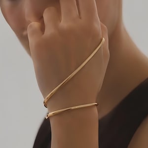 May include: A gold-toned metal bracelet featuring a continuous, smooth, and polished design that wraps around the wrist and extends diagonally across the back of the hand, creating a sleek, minimalist, and fluid silhouette with a reflective surface.