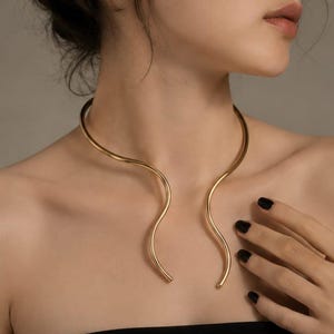 Puede incluir: Un collar tipo gargantilla de metal en tono dorado con un diseño liso y abierto, que presenta dos curvas onduladas simétricas y suaves que se extienden hacia abajo desde el cuello, creando una línea fluida alrededor de la clavícula.