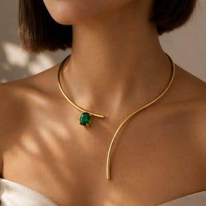 Könnte beinhalten: Eine goldfarbene, offene Halskette mit einem glatten, geschwungenen Metallband, das sich um den Hals legt. Ein Ende der Kette endet in einer polierten goldenen Spitze, während das andere Ende mit einem einzelnen, oval geschliffenen, dunkelgrünen Edelstein verziert ist.