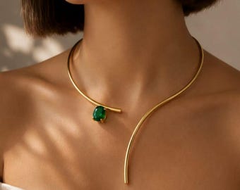 Puede incluir: Un collar abierto de tono dorado con una banda de metal lisa y curva que rodea el cuello. Un extremo del collar termina en una punta dorada pulida, mientras que el otro extremo está acentuado con una sola piedra preciosa de color verde oscuro y corte ovalado.