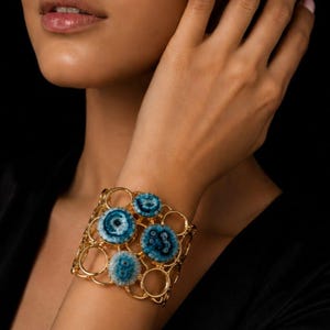 Könnte beinhalten: Ein goldfarbenes Manschettenarmband mit einem geometrischen Rahmen aus offenen Kreisen. Das Armband ist mit vier runden, blau-weißen Steinen in Achat-Optik verziert, die jeweils ein markantes konzentrisches Muster und eine texturierte Oberfläche aufweisen.